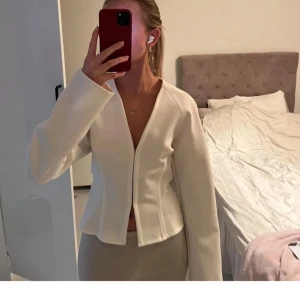 Vit figursydd kofta med v-ringning - Säljer en stilren vit kofta med djup v-ringning och lång ärm. Koftan är figursydd med markerad midja och har en minimalistisk look utan synliga knappar eller dragkedja. Perfekt för dig som gillar clean och modern stil.
