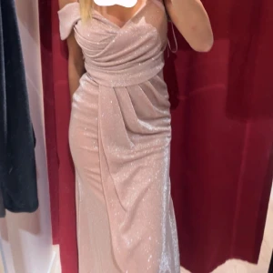 Glittrig champagnefärgad långklänning - Supersnygg långklänning i champagnefärg med glittrigt tyg och draperad överdel. Klänningen har offshoulder-ärmar och markerad midja som ger en elegant siluett. Perfekt för dig som vill sticka ut på bal eller fest!