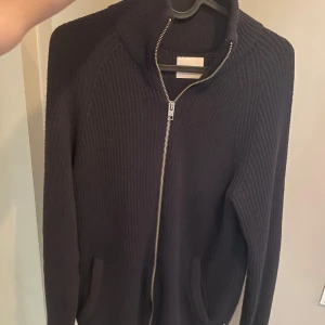 Marinblå stickad kofta från Jack & Jones - Säljer en stickad kofta från Jack & Jones i storlek S. Koftan har hel dragkedja framtill, hög krage och två fickor. Den är ribbstickad och har långa ärmar, perfekt för kyliga dagar.