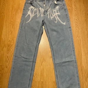 Feta jeans med vit Revenge-brodyr - Snygga jeans från Revenge med stor vit brodyrtext framtill. Klassisk femficksmodell med raka ben och normal passform. Jeansen har en cool streetwear-vibe och är tillverkade i bomull. Perfekta för dig som vill sticka ut med detaljer.