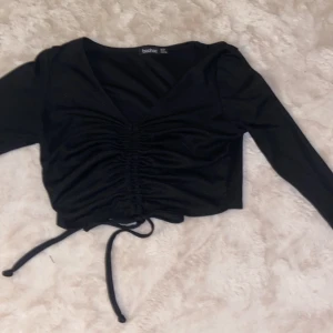 Svart croppad topp med snörning Boohoo - Säljer en svart croppad topp från Boohoo med långa ärmar och snygg v-ringning. Toppen har snörning framtill som ger en rynkad effekt och gör att du kan justera passformen. Perfekt till jeans eller kjol för en trendig look.