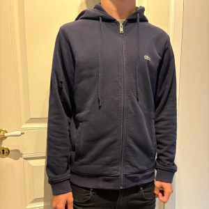 Mörkblå hoodie från Lacoste med dragkedja - Snygg mörkblå hoodie från Lacoste med dragkedja och huva med stora vita bokstäver. Mjuk fin fleece på insidan, snörning i huvan och klassisk krokodil-logga på bröstet. Perfekt för chill dagar och streetstyle. Fint material och ganska tjock, håller värme bra. Litet hål på armen