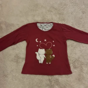 Vinröd långärmad pyjamaströja med katter - Supergullig vinröd långärmad tröja från Lindros med broderade katter på framsidan, hjärta och stjärnor. Insidan av halsen och ärmsluten har ett mönster med stjärnor och månar i grått och vinrött. Perfekt för chill dagar hemma.