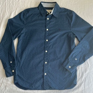 Mörkblå prickig skjorta från H&M L.O.G.G. - Mörkblå prickig skjorta från H&M L.O.G.G. med klassisk krage. Skjortan har långa ärmar, vita knappar och ett litet broderat motiv på bröstet. Tillverkad av  bomull och är i fint skick samt hel o ren. Storlek 164.