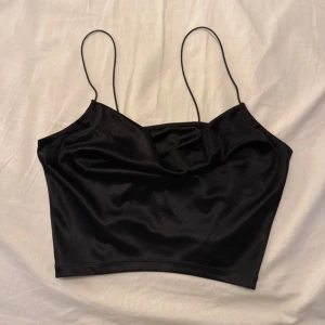 Svart croppad linnetopp med tunna band - Snygg svart croppad linnetopp med tunna axelband och lätt draperad halsringning. Materialet har en silkeslen finish som ger en cool och stilren vibe. Perfekt att matcha med jeans eller kjol för en trendig look.