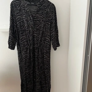 Zebramönstrad långklänning Soaked in Luxury - Säljer en svart och grå långklänning från Soaked in Luxury med zebramönster. Klänningen har trekvartsärm, v-ringad hals och snörning i midjan för en snygg passform. Materialet är lätt och luftigt, perfekt för att sticka ut med coola detaljer.