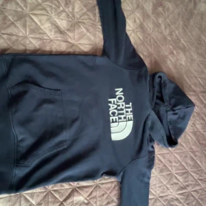 Mörkblå hoodie från The North Face - Snygg mörkblå hoodie från The North Face i juniorstorlek XL. Tröjan har stor vit logga på bröstet, huva och en klassisk magficka. Perfekt för chill dagar och streetstyle. Materialet är mjuk bomull och polyesterblandning som känns skönt mot huden. PRIS KAN DISKUTERAS PÅ ALLA VÅRA KLÄDER BARA OCH SKRIVA