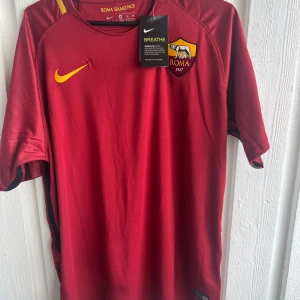 AS Roma Totti 10 matchtröja Nike - Snygg AS Roma fotbollströja med Totti 10 på ryggen, officiell Serie A-patch på ärmen och klubbmärke på bröstet. Tröjan är mörkröd med gula detaljer och tillverkad i lätt, ventilerande polyester. Perfekt för dig som älskar italiensk fotboll och legender.