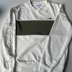Vit sweatshirt från Lacoste med logga - Säljer en clean vit sweatshirt från Lacoste i bomull med en bred olivgrön panel över bröstet och klassisk krokodillogga. Långärmad med stor LACOSTE-text längs ena ärmen. Perfekt för dig som gillar stilrena och sportiga vibes.
