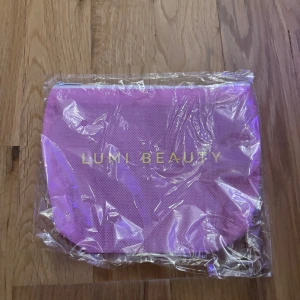 Rosa necessär från Lumi Lashes/Beauty - Snygg rosa necessär från Lumi lashes/Beauty med guldfärgad text på framsidan. Väskan har en enkel, rundad form och stängs med dragkedja upptill. Perfekt storlek för smink eller småprylar, tillverkad i syntetmaterial med meshstruktur.