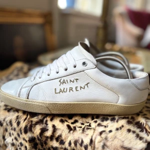 Vita Saint Laurent sneakers i skinn - Säljer ett par ÄKTA sneakers från Saint Laurent med handskriven logga på sidan. Skorna är tillverkade i Italien och har en klassisk rund tå, platt sula och snörning. Materialet är mjukt skinn och de har en stilren, minimalistisk look som passar till jeans eller dressade outfits storleken är 41,5 men passar 42 skicka är 7/10 men har mycket att ge priset kan diskuteras