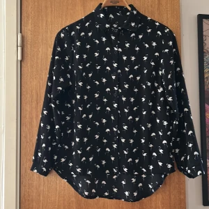 Svart skjorta med flamingomönster - Svart skjorta med vita flamingos över hela tyget. Skjortan har klassisk krage, knappar framtill och långa ärmar med knapp vid manschetten. Materialet är lätt och har en lite skrynklig struktur som ger en cool vibe. Perfekt för dig som gillar unika prints och vill sticka ut.