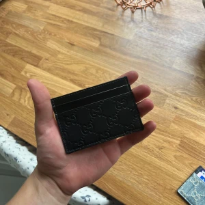 Svart Gucci plånbok  - Snygg svart kortplånbok från Gucci i äkta skinn med präglat GG-mönster. Slimmad design med plats för flera kort, perfekt att ha i fickan eller väskan. Stilren och lyxig accessoar som lyfter din look.