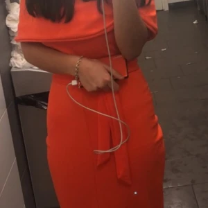 Orange offshoulder klänning med knyt - Säljer en snygg orange offshoulder klänning med knyt i midjan. Klänningen har en figurnära passform och är tillverkad i ett mjukt, strukturerat tyg. Perfekt för dig som vill sticka ut med en färgstark look och gillar trendiga detaljer.