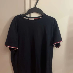 Mörkblå Moncler t-shirt slim fit - Snygg mörkblå t-shirt från Moncler i slim fit-modell. T-shirten har korta ärmar med vit och röd kant, samt Moncler-logga på vänster ärm. Tillverkad i mjuk bomull och har en klassisk rund hals. Perfekt för dig som gillar stilrena och exklusiva plagg. Priset är inte hugget i sten, så skicka ett rimligt pris!