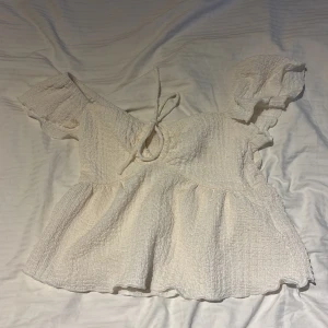 Vit blus med puffärm från DIVIDED - Supersöt vit blus från DIVIDED i storlek XXS. Den har puffiga korta ärmar, knytning fram och en lätt volang nedtill. Materialet är krinklat och känns mjukt mot huden.