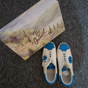 Blå och vita sneakers från The Gallerist strl 41 - Säljer ett par snygga sneakers från The Gallerist i vitt skinn med blå detaljer och klubbmönster på sidan och hälen. Skorna har snörning, rund tå och platt sula. Insidan har färgglad prickig innersula och text. Perfekt för dig som gillar unik design! Bara använda 2-3 gånger! Här av dig vid minsta fundering! 😁