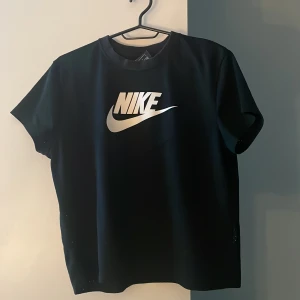 Svart Nike t-shirt med meshdetaljer - Svart t-shirt från Nike med stor vit logga på bröstet. Modellen är kortärmad och har meshpaneler med små hål i hela tröjan  och sidor för extra ventilation. Materialet känns lätt och sportigt, perfekt för dig som gillar en clean och modern look.