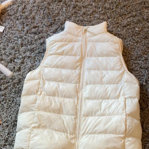 Vit dunväst från Uniqlo - Säljer en fräsch vit dunväst från Uniqlo med ståkrage och dragkedja framtill. Västen har quiltad design och är lätt vadderad, perfekt för lager-på-lager. Passformen är normal och materialet känns mjukt. Enkel och clean look som funkar till jeans eller hoodie. Den passar xs-s