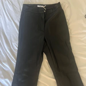 Svarta skinnbyxor från NA-KD - Snygga svarta kostymbyxor från NA-KD med rak passform och klassisk midja. Byxorna har en stilren design med knapp och dragkedja framtill. Materialet känns mjukt och har en lätt glansig finish som ger en modern vibe. Perfekta att styla med sneakers eller boots.