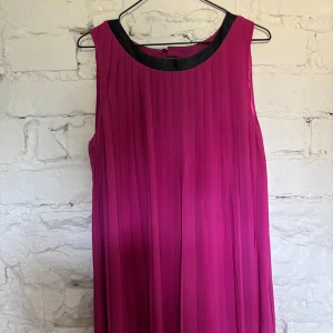 Plisserad cerise klänning med svart kant - Säljer en snygg plisserad klänning i cerise med svart kant vid halsen. Klänningen är ärmlös och har en rak, luftig passform som ger en cool och modern vibe. Perfekt för dig som gillar färg och vill sticka ut lite extra.