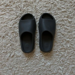 YEEZY SLIDES- mörkgrå - Svarta Adidas Yeezy Slides med minimalistisk design och präglad Adidas-logga på fotbädden. Slidesen har en chunky, räfflad sula som ger cool vibe och extra komfort. Tillverkade i syntetmaterial och passar perfekt för chill dagar eller när du vill ha en clean look. 43 men passar mindre- 41,5-42 isch.