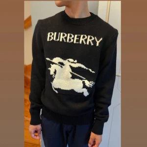 Svart stickad tröja från Burberry, S  - Snygg svart stickad tröja från Burberry med ett stort vitt logotyptryck och en riddare på framsidan. Storlek S. Oanvänd. Färg: Svart. 