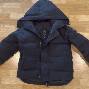 Svart dunjacka från Canada Goose - Säljer en svart dunjacka från Canada Goose med huva och snyggt broderat märke på ärmen. Jackan har dragkedja och knappar framtill, två sidofickor och en innerficka. Perfekt för kalla vinterdagar och har en clean, puffad look som är riktigt stilren.