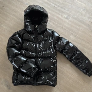 Svart Colmar pufferjacka med huva - Säljer en svart, glansig pufferjacka från Colmar med huva och dragkedja framtill. Jackan har ett stilrent, quiltat mönster och logotyp på ärmen. Perfekt för dig som vill ha en varm och snygg jacka med modern vibe. Passar dig som gillar streetstyle och vill sticka ut. Storlek 16 Y (172cm)
