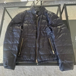 Mörkblå pufferjacka från Prada - Snygg pufferjacka från Prada med quiltad design och glansig finish. Jackan har ståkrage, två stora fickor med dragkedja och ikonisk Prada-logga på ärmen. Perfekt för dig som vill ha en stilren och exklusiv look under kyliga dagar.