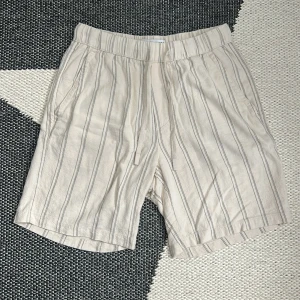 Beige randiga shorts från Zara - Snygga beige shorts från Zara med svarta vertikala ränder och elastisk midja med snörning. De har en avslappnad passform, sidofickor och en bakficka. Materialet känns lätt och luftigt, perfekt för varma dagar.