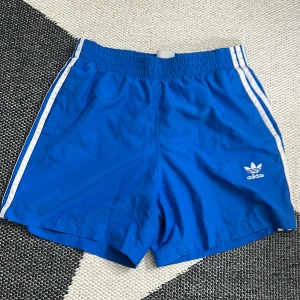 Blå Adidas shorts med vita ränder - Snygga blå shorts från Adidas med klassiska vita ränder längs sidorna och Adidas-logga framtill. De har elastisk midja och är gjorda i lätt polyester med meshfoder. Perfekta för träning, bada eller chill dagar. Sportig vibe och skön passform.