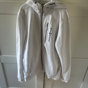 Vit hoodie från Sail Racing med dragkedja 160 - Säljer en fräsch vit hoodie från Sail Racing med hel dragkedja framtill och broderad logga på bröstet. Tröjan har huva, långärmade ärmar och en praktisk bröstficka med dragkedja. Perfekt för chill dagar och snygg till jeans eller joggers. Storlek 160