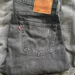 Levi's 501 svarta jeans straight fit - Svarta Levi's 501 jeans med klassisk straight fit och knappgylf. Jeansen har fem fickor, silverfärgade och kopparfärgade nitar samt den ikoniska röda Levi's-taggen vid bakfickan. Materialet är slitstarkt denim i bomull och färgen är mörkgrå/svart. Nästintill oanvända. 9/10 skick. Storlek 32 i bredd och 34 i längd. Säljer för endast 399kr nypris 1000kr+