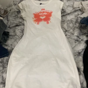 Vit kortklänning med tryck från H&M Divided - Säljer en vit kortklänning från H&M Divided i storlek XS. Klänningen har ett coolt orange tryck med texten 'The Utopia Life' och en fjäril med hjärta på bröstet. Modellen är figurnära med korta ärmar och rund hals. Perfekt för dig som gillar streetstyle och grafiska prints.