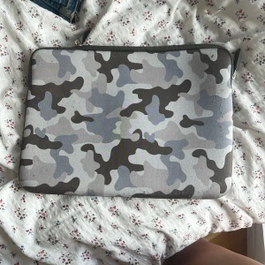 Camouflage datorväska med dragkedja - Snygg datorväska med grått och svart camouflage-mönster. Väskan har en praktisk dragkedja upptill och är fodrad på insidan för extra skydd. Perfekt för att bära med sig laptopen till skolan eller jobbet. typ aldrig använt