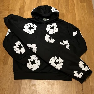 Svart hoodie från Denim Tears  - Svart hoodie från Denim Tears med stora vita blommönster över hela tröjan. Tröjan har huva och långa ärmar med muddar. Materialet är mjuk bomull och passformen är normal. Perfekt för dig som gillar streetwear och vill sticka ut med en unik design.