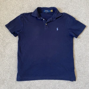 Marinblå Polo Ralph Lauren t-shirt - Snygg marinblå piké från Polo Ralph Lauren i slim fit-modell. Klassisk pikékrage med två knappar och ikonisk broderad logga på bröstet. Tillverkad i mjuk bomull som är skön mot huden. Den är i väldigt bra skick. Skriv gärna vid frågor! 🌟