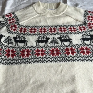 Jultröja med renar från Ellos - Mysig stickad jultröja från Ellos i storlek 42/44. Tröjan är vit med klassiskt mönster i svart och rött, med renar, granar och snöflingor. Rund hals och långa ärmar med ribbade muddar. Perfekt för vinter och julmys!