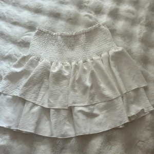 Vit volangkjol med smock - Supersöt vit kortkjol med bred smock i midjan och dubbla volanger. Kjolen har en luftig och flowy känsla, perfekt för varma dagar. Materialet känns lätt och har en fin struktur. Passar dig som gillar romantisk och feminin stil.