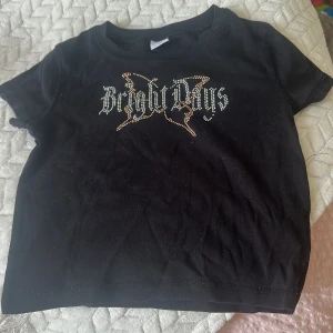 Svart topp med strass från Lindex - Cool svart topp från Lindex med korta ärmar och rund hals. Framsidan har ett snyggt strassmotiv med texten 'Bright Days' och en fjäril i silver och guld. Materialet är mjuk bomull som känns skönt mot huden. Perfekt för dig som gillar lite extra bling i vardagen.