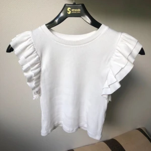 Vit volangtopp från Zara - Supersöt vit topp från Zara med breda volangärmar som ger en extra touch. Modellen är croppad och har en rund halsringning. Materialet är mjukt och ribbat, perfekt för dig som gillar en clean och fräsch look med lite extra detaljer.