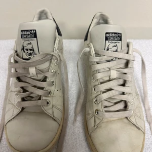 Adidas Stan Smith vita sneakers - Säljer ett par klassiska Adidas Stan Smith sneakers i vitt med mörkblå häl och logga. Skorna har rund tå, platt sula och snörning framtill. Materialet är skinn och de har perforerade detaljer på sidorna. Perfekt för dig som gillar retrostil och ikoniska modeller.