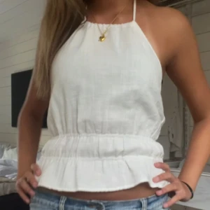 Vit linnetopp med volangkant öppen rygg - Fräsch vit linnetopp med halterneck och volangkant nedtill. Toppen är ärmlös och har en croppad passform som sitter snyggt till jeans eller shorts. Perfekt för varma dagar och har en lätt, luftig känsla tack vare materialet.