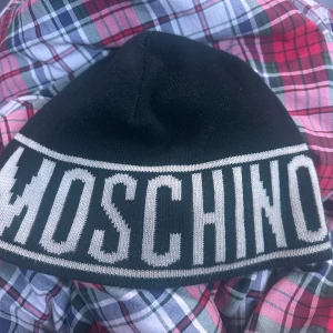 Svart Moschino mössa med logga - Svart mössa från Moschino med stor vit logotyp runt hela kanten och Milano-text. Snygg, stilren design som sticker ut. Tillverkad i 50% ull och 50% akryl, perfekt för kalla dagar. Passar dig som gillar exklusiva streetwear-vibbar.