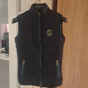 Mörkblå dunväst från Jacson - Snygg mörkblå dunväst från Jacson med guldfärgad dragkedja och två fickor med detaljer i turkos. Västen har ett broderat emblem på bröstet och hög krage. Perfekt för dig som gillar ridning eller vill ha en sportig look.