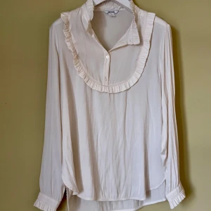 Vit volangskjorta från Monki - Säljer en vit skjorta från Monki i storlek L med snygga volangdetaljer runt kragen och knäppning framtill. Skjortan har lång ärm med volang vid manschetten och är tillverkad i ett mjukt, lätt material som ger en skön känsla. 