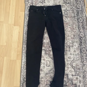Svarta jeans från J.Lindeberg - Svarta jeans från J.Lindeberg med klassisk femficksdesign och silverfärgad knapp. Slimmad passform med skinny stil och dragkedja i gylfen. Snyggt JL-logo på myntfickan och läderpatch med logga baktill. Perfekta för en clean och stilren look.