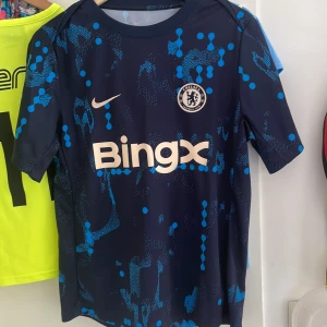 Äkta Chelsea FC blå träningströja Nike M - Chelsea FC träningströja i storlek M från Nike med Dri-Fit-material. Tröjan är marinblå med ett unikt blått mönster och har korta ärmar. Tryck med BingX och Infinite Athlete på bröst och ärm. Perfekt för dig som vill ha en snygg och modern fotbollströja.
