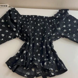 Svart blommig blus från Kappahl - Supersöt svart blus från Kappahl med vita blommor och smockad midja. Toppen har offshoulder-design och puffiga korta ärmar med volangkant. Materialet är mjukt och stretchigt, perfekt för dig som gillar romantisk stil och vill sticka ut lite extra.